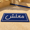 MAALESH VIBES die-cut rug — the Arabic word معلش in white on a navy street-sign plaque, cut to the rectangle with black edge binding