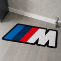 M Performance die-cut rug — BMW M badge with blue, navy and red stripes and a white M, cut to the badge shape with black edge binding