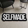 Self-Made Hustle die-cut rug — $ELFMADE in heavy white block letters on black, cut to the shape of the word with black edge binding