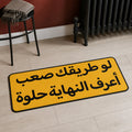 Worth The Road die-cut rug — the Arabic line لو طريقك صعب أعرف النهاية حلوة in black on an amber plaque, cut to the sign shape with black edge binding