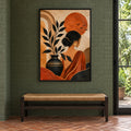 Front view of The Terracotta Muse in a black frame above a woven bench, showing a woman, leafy branches and a terracotta sun.